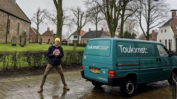 Toukomstcollega Thijs staat vrolijk bij de Toukomstbus met een Toukomstmuts op zijn hoofd.