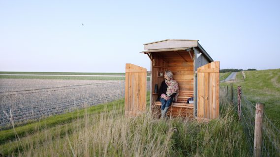Vrouw in een klein houten hutje tussen lang uitgestrekte akkers.